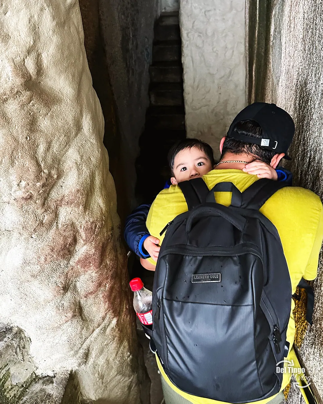 Padre cargando a Sebastián durante la bajada por las escaleras estrechas dentro de la Piedra del Peñol, con mochila negra y el muro de roca a los lados