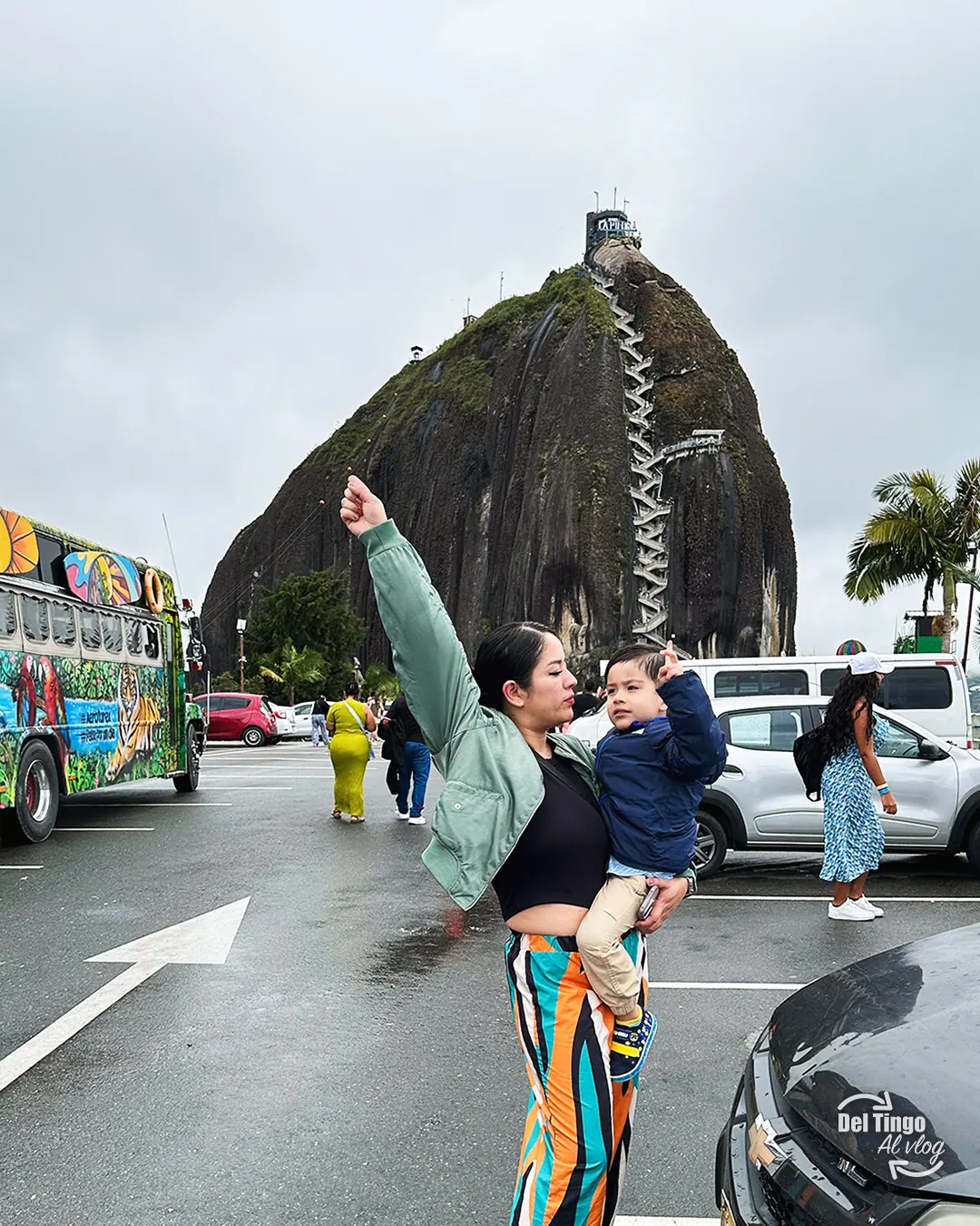 Mamá cargando a Sebastián y señalando hacia la cima de la Piedra del Peñol al fondo, con una chiva colombiana decorada estacionada a la izquierda