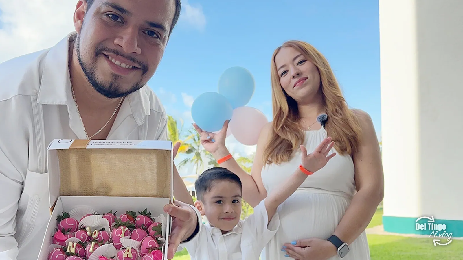 Familia en Cancún durante el gender reveal: papá sosteniendo una caja con fresas decoradas, mamá embarazada con globos rosas y azules y su hijo en medio