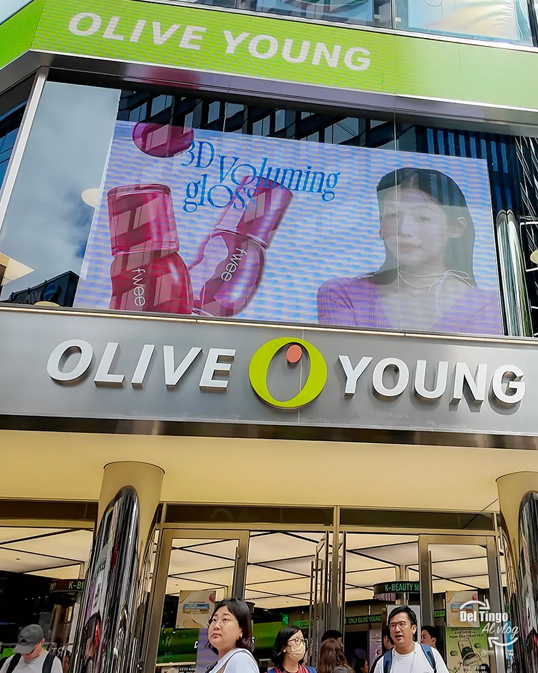 Entrada vertical de la tienda Olive Young en Myeongdong, Seúl, con doble letrero corporativo, pantalla publicitaria de productos de K-beauty y varias clientas saliendo del local