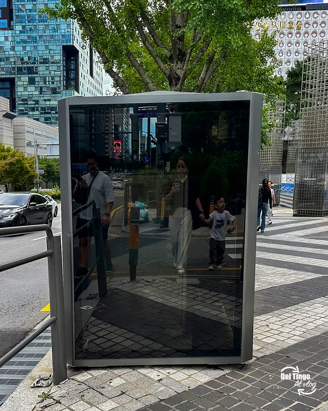 Familia reflejada en un panel-espejo gigante en una calle de Gangnam, Seúl, con un niño tomado de la mano y rascacielos al fondo